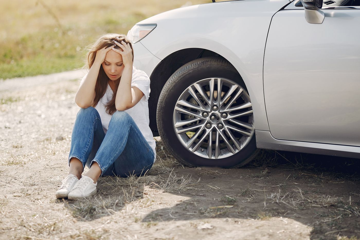 A driver sitting on the ground beside her car with her hands on her head